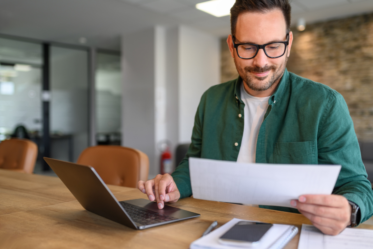 Homem concentrado em frente a um notebook, com uma mão no teclado e segurando um documento na outra, representando a tarefa de renovação de um e-CNPJ A1 com validade estendida.
