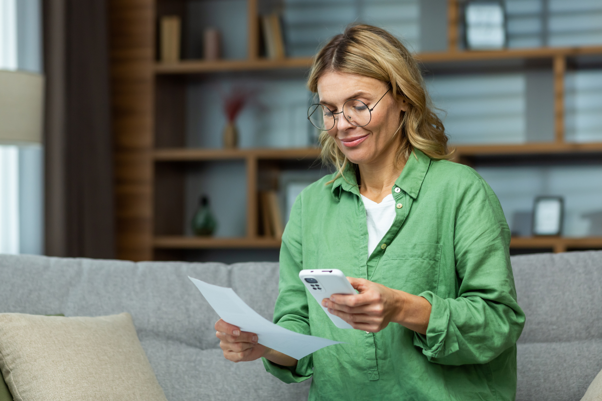 Empresária trabalhando em home office, sentada no sofá com celular e documento em mãos, analisando informações sobre certificado digital.