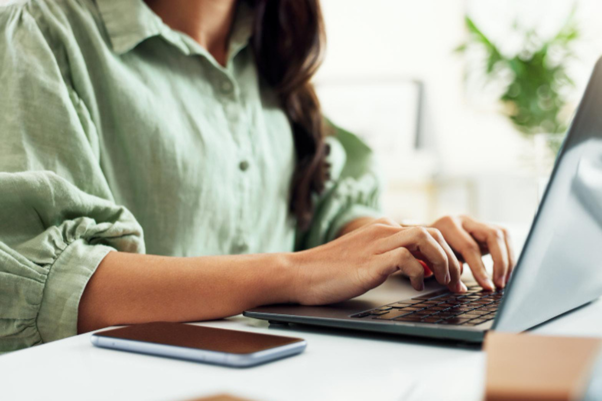 Mulher sentada usando notebook sobre uma mesa com um celular ao lado em ambiente de escritório representando: CBS e IBS: o que são, quando começam e como afetam seu dia a dia.