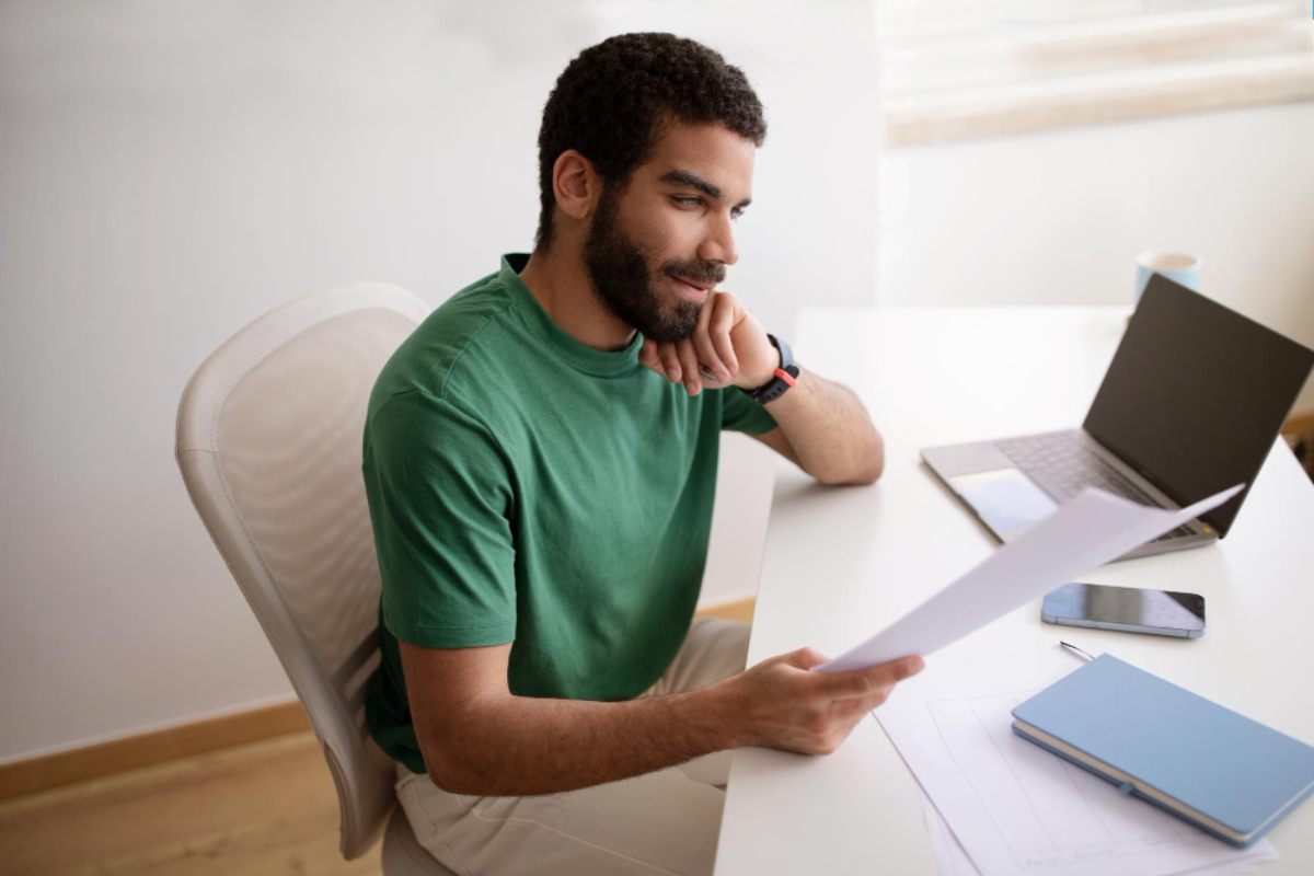 Homem em ambiente de escritório sentado com uma folha ( documento) em uma das mãos em frente ao notebook sobre uma mesa com celular e agenda ao lado representando: Reforma tributária para o contador: cronograma, rotinas e entregáveis por fase.