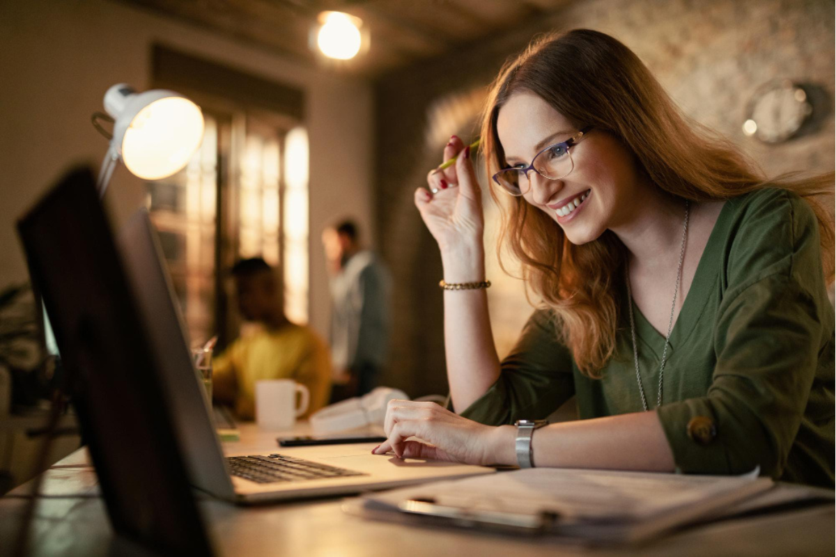 Mulher sorrindo sentada com canta na mão direita em frente a um notebook sobre uma mesa com uma prancheta com papéis do lado esquerdo e uma xícara do lado direito, mais `a sua direita dois homens, um em frente a uma janela e o outro também usando um notebook, todos em ambiente de escritório representando: Os aspectos contábeis da Reforma Tributária.