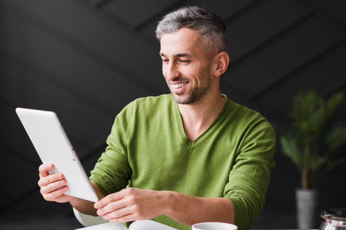 Homem sorridente em ambiente de escritório sentado em uma mesa com livro aberto e uma xícara de café ao lado usando um tablet representando: O que é o carimbo do tempo e por que ele importa em documentos assinados digitalmente.