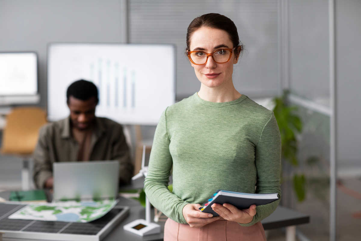 Mulher em pé com pasta de documentos em mãos em ambiente de escritório com homem ao fundo sentado fazendo uso de um notebook sobre uma mesa representando: Reforma Tributária: todas as respostas que você precisa.