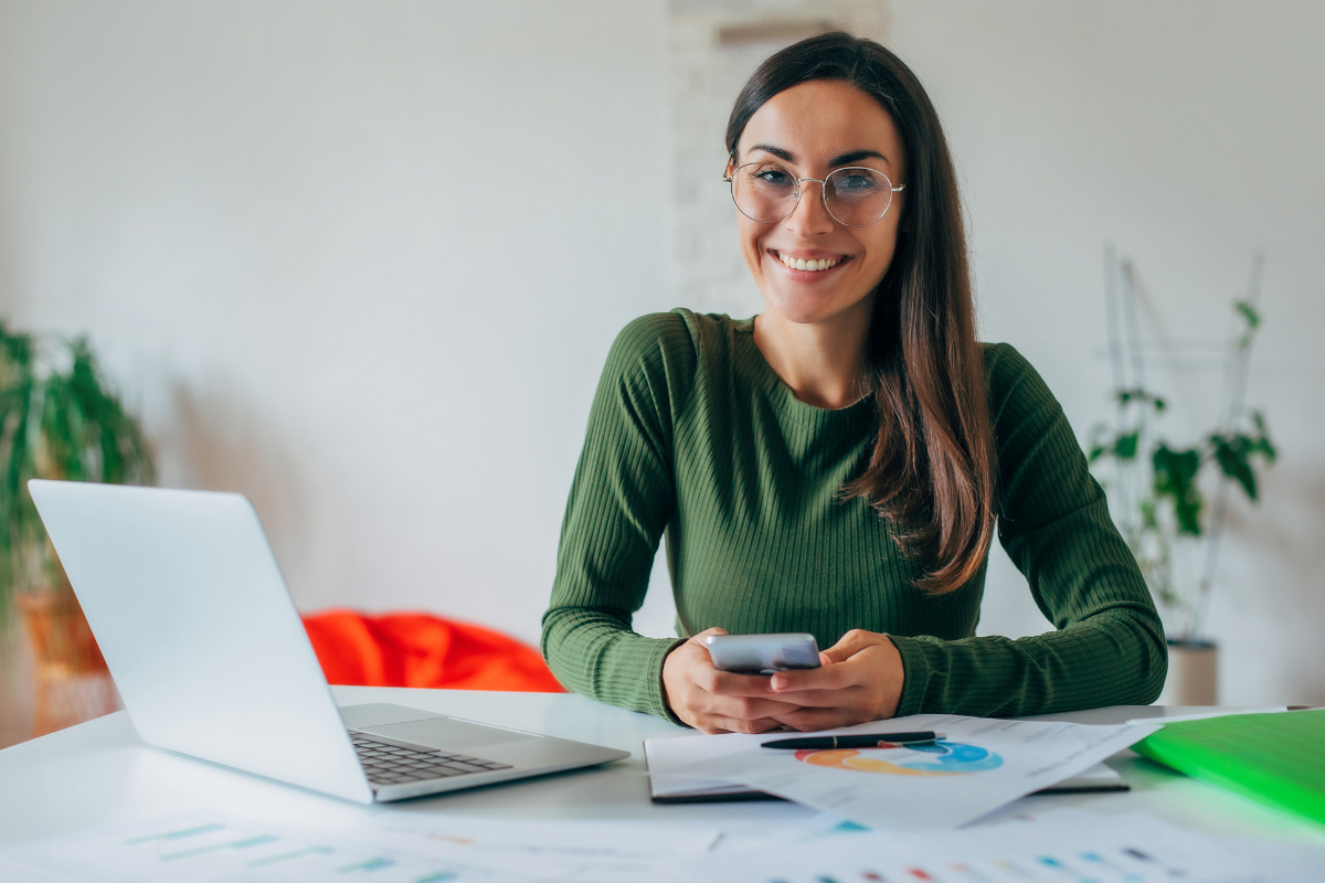 Empresária em ambiente de escritório sentada sorridente em frente a um notebook com celular em mãos em uso, com papéis, documentos e caneta sobre a mesa representando: Tudo que você precisa saber sobre IVA dual.