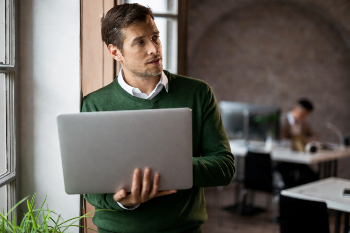 Homem usando notebook em mãos e de pé em ambiente de escritório conversando com colegas representando: Obrigações acessórias que exigem certificado.