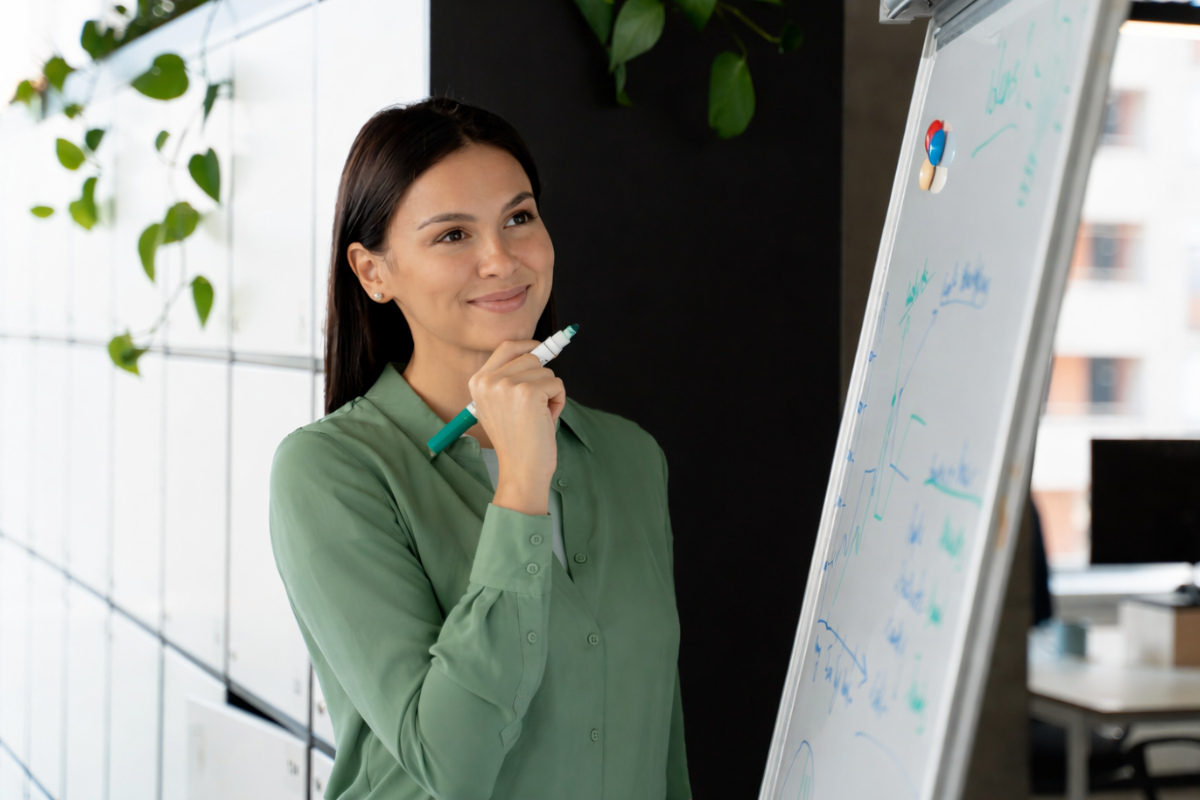 Mulher de pé em frente a um quadro negro com caneta em mãos analisando o que acabara de escrever no quadro em ambiente de escritório representando: Precificação e Reforma Tributária: quando é necessário reavaliar?