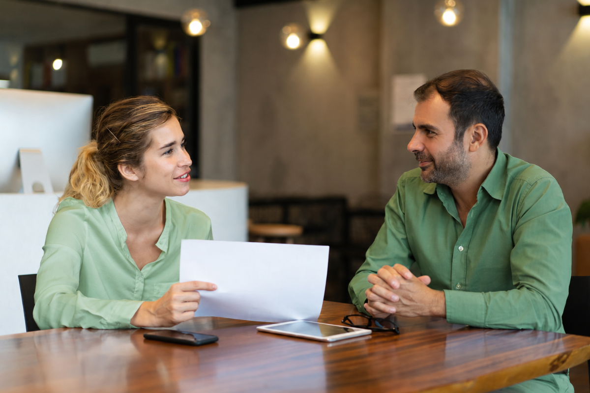 Um homem e uma mulher conversando sobre projetos em uma mesa de escritório com papeis em mãos representando: Como sair do modelo operacional e ir para o consultivo.