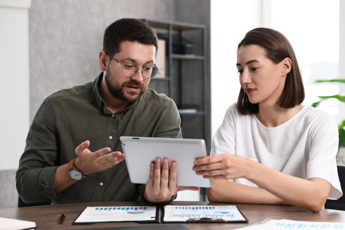 Homem e mulher conversando em uma mesa de escritório com tablet em mãos e com documentos, prancheta e sobre a mesa representando: Como fidelizar clientes contábeis.