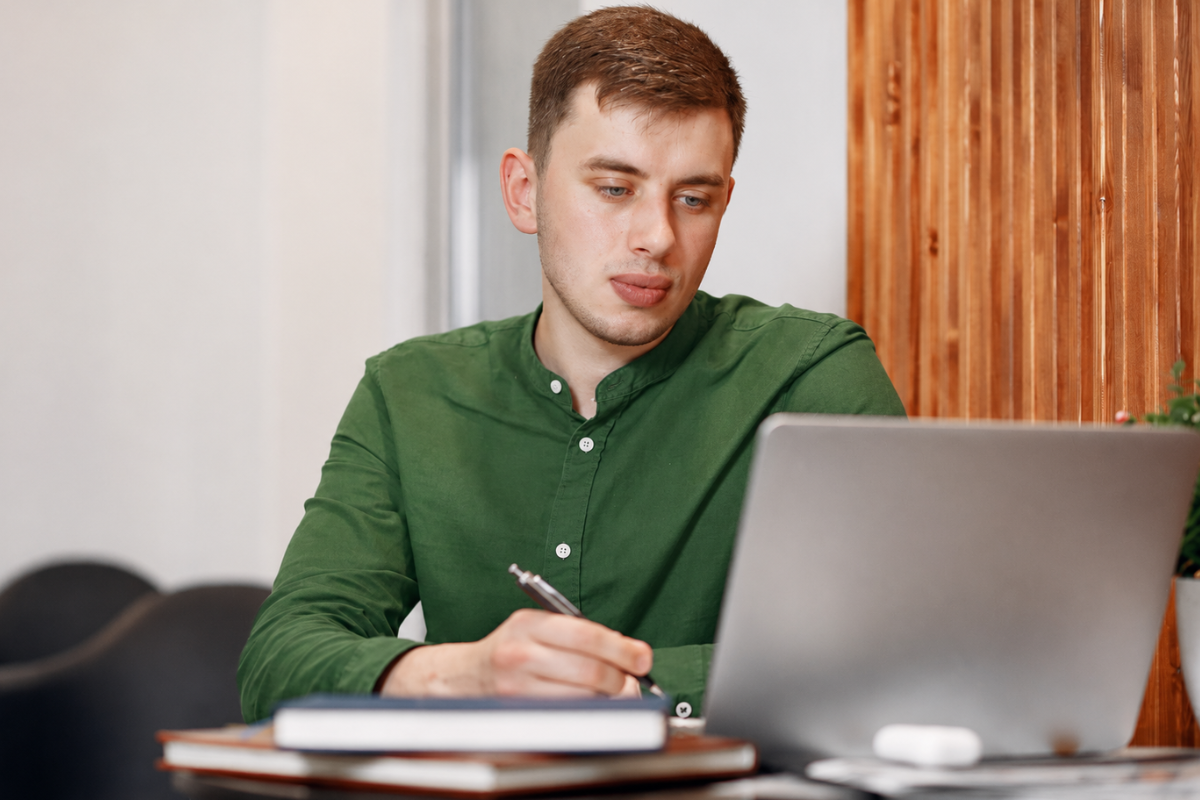 Jovem empresário trabalhando em um notebook e fazendo anotações em uma agenda em ambiente de escritório representando: SPED e certificado digital: qual a relação?