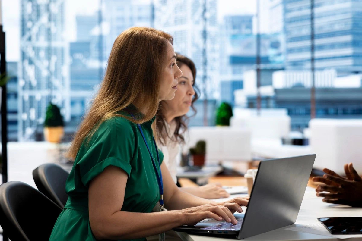 Duas empresárias trabalhando em seus notebooks sobre uma mesa em ambiente de escritório sobre gestão de acessos digitais na microempresa: certificados, senhas e permissões em um só lugar.